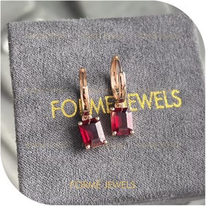 May include: Rose gold hoop earrings, each featuring a rectangular, deep red gemstone. The earrings are presented on a grey pouch with the gold text "FORME JEWELS". The gemstones have a rich, saturated colour.
