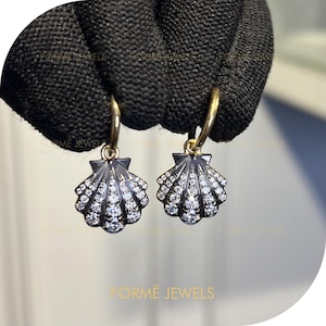 May include: A pair of gold hoop earrings featuring seashell pendants. The pendants are black with a fan design, set with small, sparkling white stones. The earrings are held by a gloved hand.
