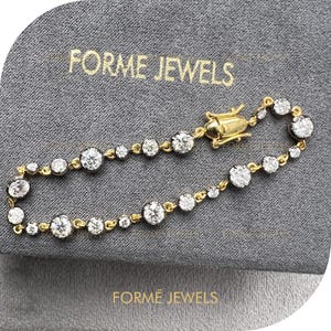 May include: A gold bracelet with multiple clear, round gemstones. The bracelet is set on a gray surface with the text "FORME JEWELS" printed in gold.