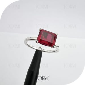 Peut inclure: Une bague en argent avec une grande pierre rouge de forme carrée. La bague est sur un support noir.