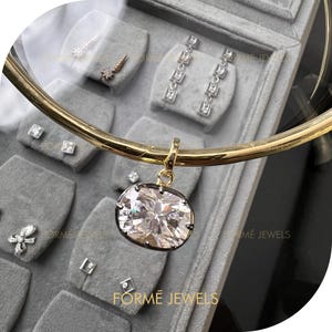 May include: A gold-coloured necklace with a large, square-cut pendant featuring a sparkling, clear gemstone. The pendant is set in a dark metal frame. Other jewellery pieces, including earrings and studs, are displayed in a grey jewellery box.