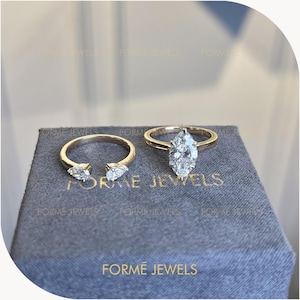 May include: Two gold rings resting on a gray box with the text FORME JEWELS. One ring is an open band featuring two pear-cut diamonds at the ends. The second ring is a solid band set with a single large marquise-cut diamond in the center.