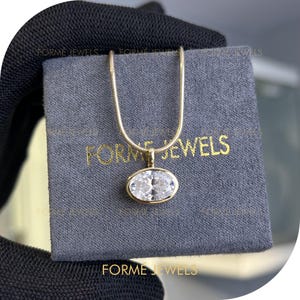 Puede incluir: Un collar dorado con un colgante ovalado de piedra preciosa transparente. El colgante está engastado en un bisel dorado. El collar descansa sobre una tela gris con el texto "FORME JEWELS" en dorado. El collar se muestra sobre un fondo oscuro.