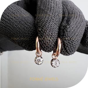 May include: Rose gold hoop earrings with a small, round, clear gemstone pendant. The earrings are displayed against a dark grey textured background. The brand name "FORME JEWELS" is visible in gold lettering.