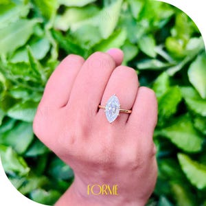 Puede incluir: Un anillo de oro con un gran diamante claro de forma marquesa. El anillo está en una mano con el fondo desenfocado.