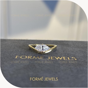 Op de afbeelding: Een gouden ring met een heldere, marquise-geslepen edelsteen in een V-vormige band. De ring ligt op een donkergrijs oppervlak met de tekst FORME JEWELS in gouden letters gedrukt. Het ontwerp toont een gepolijste metalen afwerking en een facetsteen.