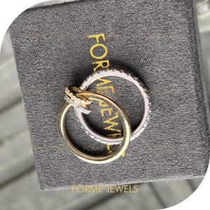 May include: Two interlocking rings resting on a dark gray textured box with the gold text FORME JEWELS printed on it. One ring is a smooth gold band, while the other is a silver-toned band encrusted with small clear stones along its entire outer surface.