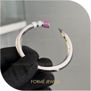 以下が含まれることがあります： 曲線的なデザインのシルバーカラーのブレスレット。ブレスレットには、白とピンクの宝石が付いたセクションがあります。「FORME JEWELS」の文字が画像の下部に表示されています。