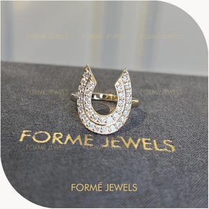 May include: A gold ring featuring a horseshoe-shaped centerpiece adorned with numerous small, clear, sparkling stones. The text FORME JEWELS is printed in gold lettering on the dark gray surface beneath the ring, repeating the brand name.