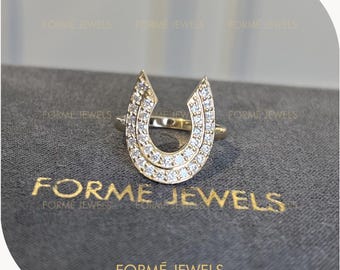 May include: A gold ring featuring a horseshoe-shaped centerpiece adorned with numerous small, clear, sparkling stones. The text FORME JEWELS is printed in gold lettering on the dark gray surface beneath the ring, repeating the brand name.