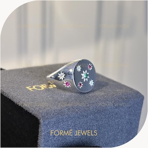 May include: A silver signet ring with a dark gray face, adorned with star-shaped gemstones in white, pink, and green. The ring is set on a gray fabric surface, with the brand name "FORME JEWELS" visible.