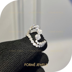 Può includere: Un anello d'argento con numerose pietre preziose rotonde e trasparenti. L'anello presenta un design curvo unico, con pietre più grandi raggruppate nella parte superiore. Il marchio "FORME JEWELS" è visibile in basso nell'immagine.