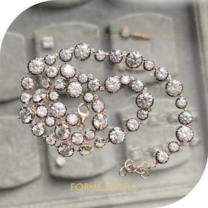 Puede incluir: Un collar de oro rosa con numerosas piedras preciosas redondas y transparentes. Las piedras están engastadas en un metal oscuro, creando un contraste con la cadena de oro rosa. El collar se exhibe sobre una superficie reflectante. La marca "FORME JEWELS" es visible.