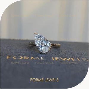 May include: A pear-shaped diamond ring featuring a clear, faceted stone set on a thin, polished gold band. The text FORME JEWELS is visible in gold lettering on the dark, textured surface beneath the ring, repeated multiple times in the background.