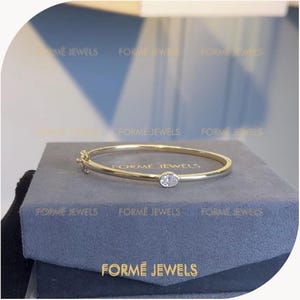 May include: A gold bangle bracelet featuring a single oval-shaped clear stone set in a bezel setting. The bracelet includes a secure hinged clasp mechanism on the side. The text FORME JEWELS is visible on the surface of the box beneath the bracelet.