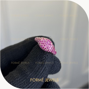 May include: A signet ring featuring a rounded top surface densely encrusted with numerous small, vibrant pink gemstones. The metal band appears to be a metallic tone, and the overall design is compact and rounded, showcasing a pavé-style setting.