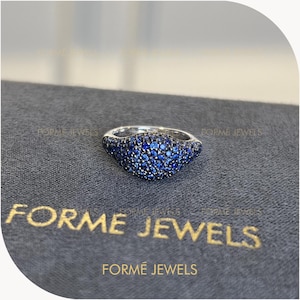Op de afbeelding: Een zilverkleurige ring met een afgerond bovenoppervlak dat dicht bezet is met talloze kleine, levendige blauwe stenen. De ring bevindt zich boven de tekst FORME JEWELS, die in gouden letters op een donkergrijs gestructureerd oppervlak staat.