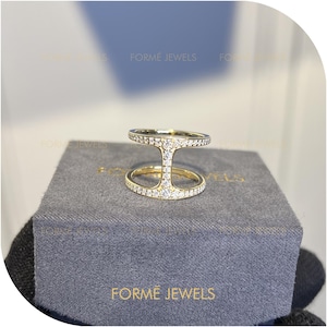 May include: A gold-colored ring with a unique open design, featuring two parallel bands connected by a vertical bar. The ring is encrusted with small, sparkling gemstones. The ring is displayed on a gray box with the text "FORMÉ JEWELS".
