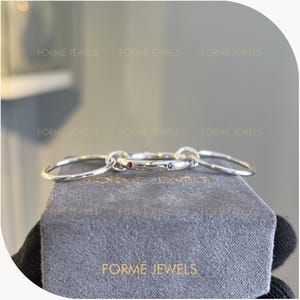 May include: A silver ring set with three connected rings, each with a polished finish. Two rings feature small gemstones, one red and one purple. The rings rest on a gray velvet surface with the text "FORME JEWELS".