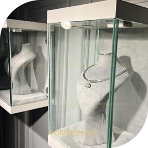May include: A silver necklace with a round pendant is displayed on a grey bust inside a glass display case. The display case is white with the text "FORME JEWELS" in gold lettering. Another display case is visible in the background.