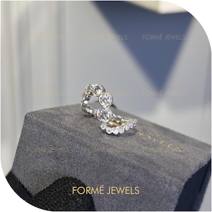 May include: A silver ring with multiple clear gemstones. The ring has a unique design with three larger stones and a band of smaller stones. The ring is displayed on a gray surface, with the brand name "FORME JEWELS" visible.