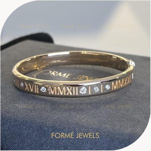 Op de afbeelding: Een goudkleurige armband met Romeinse cijfers en kleine, heldere edelstenen. De armband heeft de tekst "FORME JEWELS" en wordt gepresenteerd op een grijze ondergrond. Het is een sieraad.