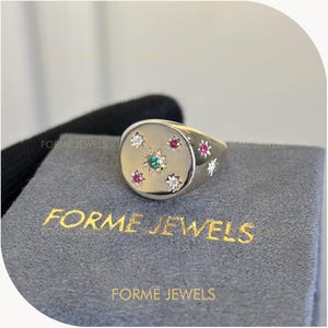 May include: A gold signet ring featuring a central star-set green gemstone surrounded by smaller star-set white and pink gemstones. The ring rests on a grey fabric surface with the gold text FORME JEWELS printed on the front of the display box.