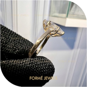 Könnte beinhalten: Ein goldener Ring mit einem großen, klaren, marquiseförmigen Edelstein. Der Ring hat ein filigranes Banddesign, und der Edelstein wird von einer detaillierten Fassung gehalten. Der Ring wird vor einem dunklen Hintergrund gezeigt. Der Text "FORMÉ JEWELS" ist sichtbar.