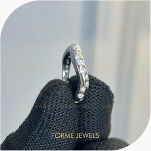 Puede incluir: Un anillo curvo de tono plateado que presenta una serie de piedras redondas transparentes incrustadas a lo largo de su superficie exterior. La banda tiene un acabado metálico pulido y muestra el texto FORME JEWELS, repetido tenuemente al fondo.