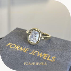 May include: A gold ring featuring a bezel-set, rectangular radiant-cut clear gemstone. The ring is positioned on a grey fabric surface with the gold-colored text FORME JEWELS printed on the fabric below the ring.