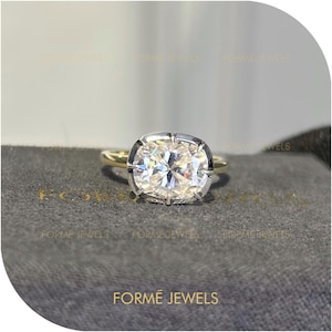 May include: A diamond ring featuring a large, square-cut diamond. The ring has a gold band and a silver-coloured setting. The ring is displayed on a grey surface. The text "FORME JEWELS" is visible in the image.