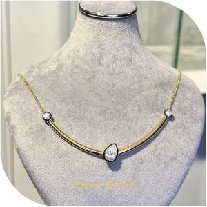 May include: A gold necklace with a curved bar design, featuring three clear gemstones. The central stone is pear-shaped, and the other two are round. The necklace is displayed on a grey mannequin.