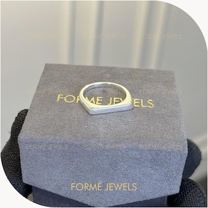 May include: A silver ring with a rectangular top and a rounded band sits on a gray box. The box has the words "FORMÉ JEWELS" printed in gold. The ring is a modern, minimalist design.