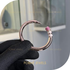 Può includere: Un braccialetto in oro rosa dal design curvo. Il braccialetto presenta una sezione ornata di pietre preziose rosa e cristalli trasparenti. Il testo "FORME JEWELS" è visibile nell'immagine.