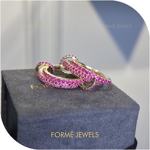 Könnte beinhalten: Zwei goldfarbene Ringe mit leuchtend rosa Edelsteinen. Die Ringe sind ineinander verschlungen und bilden ein durchgehendes Design. Sie werden auf einer dunkelgrauen Schachtel mit dem Text "FORMÉ JEWELS" präsentiert.