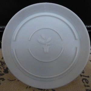 Peut inclure: Une soucoupe en plastique blanche avec un bord surélevé et un petit motif de plante au centre. Le motif représente une plante en pot avec deux feuilles.