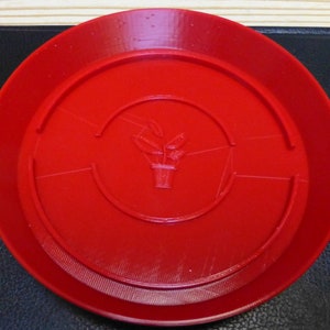 Peut inclure: Une soucoupe à plantes en plastique rouge avec un motif en relief d'une plante en pot au centre.