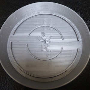 Può includere: Un sottovaso per piante color argento con bordo rialzato e un disegno centrale in rilievo. Il disegno presenta una pianta in vaso con foglie e un motivo a linee curve. Il sottovaso è progettato per raccogliere l'acqua e proteggere le superfici.