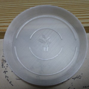 May include: A clear plastic plant saucer with a raised rim and a small plant icon in the centre.