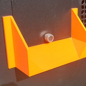 May include: A bright orange plastic shelf with a silver knob attached to the side. The shelf is mounted on a black surface.