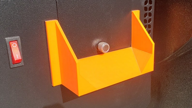 May include: A bright orange plastic shelf with a silver knob attached to the side. The shelf is mounted on a black surface.