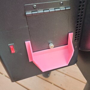 May include: A black smoker with a pink plastic tray attached to the side. The tray is designed to hold small items like grilling tools or spices.