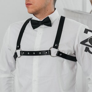 May include: A white long-sleeved shirt with a black bow tie and a black leather harness with silver buckles and rings. The harness has a studded design.