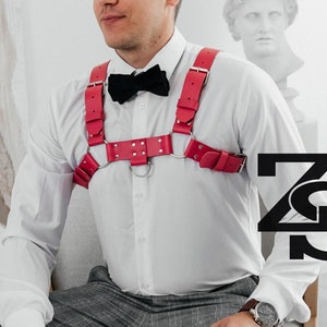 May include: A person wearing a white shirt with a black bow tie and a red leather harness with silver accents. The harness has a ring in the center and is decorated with small studs. The person is sitting on a chair with their hands in their lap. The background is a white wall with a painting on it.