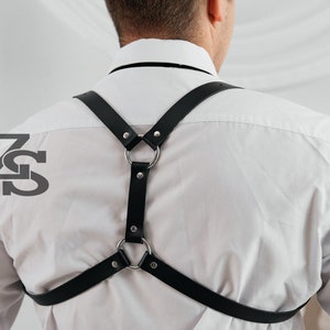 May include: A black leather harness with silver metal rings. The harness is worn over a white button-down shirt.