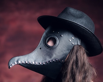May include: A person wearing a black leather plague doctor mask with a long beak and a black hat. The mask has metal rivets and a round eye hole. The person has long dark hair.