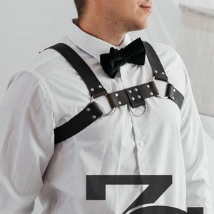 May include: A person wearing a white button-down shirt with a black bow tie and a black leather harness with silver hardware. The harness has a ring in the center and is secured with buckles. The person is also wearing a black belt and gray plaid pants.