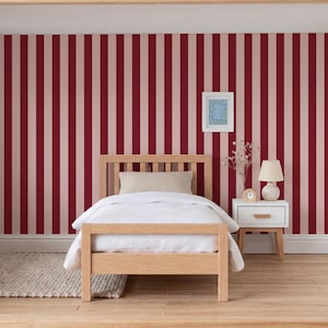 Op de afbeelding: Een miniatuur slaapkamer scène met een licht houten bedframe, wit beddengoed en een beige kussen. De muren hebben verticale strepen in bordeauxrood en lichtroze. Een kleine witte nachtkastje bevat een lamp en een klok.