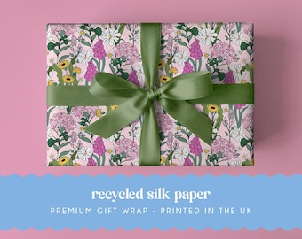 May include: A rectangular gift box wrapped in light pink paper featuring a repeating botanical pattern of white daisies, yellow flowers, and clusters of pink blossoms. The box is tied with a wide, dark olive green satin ribbon bow.