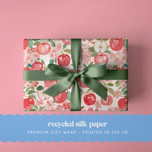 May include: A rectangular gift box wrapped in white paper featuring a repeating pattern of red apples, green leaves, and light pink blossoms. A wide, dark green satin ribbon is tied in a bow around the center of the box.